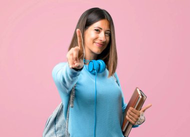 Mavi kazak ve kulaklık gösteren ve pembe arka planda en iyi işareti bir parmak kaldırma ile genç öğrenci kız