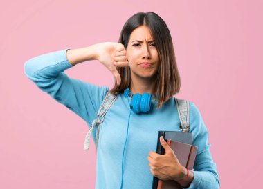 Mavi kazak ve kulaklık ile genç öğrenci kız olumsuz ifade ile başparmak aşağı işareti gösteren. Pembe arka planda hüzünlü ifade