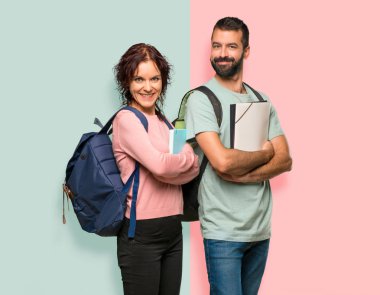 Renkli duvarda gülümseyerek kollarını yan pozisyonda geçen sırt çantaları ve kitapları olan iki öğrenci