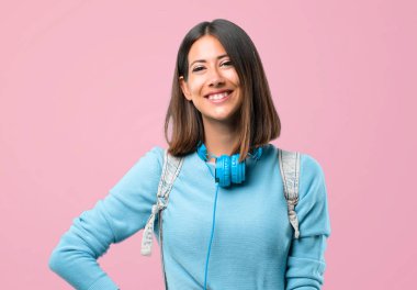 Mavi kazak ve kulaklık ile genç öğrenci kız kalça kolları ile poz ve pembe arka plan üzerinde gülümseyerek