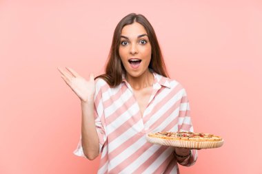 Genç kadın şok yüz ifadesi ile izole pembe duvar üzerinde bir pizza tutan