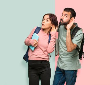 Renkli duvarda kulak elini koyarak bir şeyler dinleyerek sırt çantaları ve kitapları olan iki öğrenci