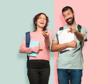 Sırt çantaları ve kitapları olan iki öğrenci birini parmağıyla işaret ediyor ve renkli duvarda çok gülüyorlar.