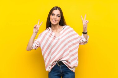 İzole edilmiş sarı arka planda genç bir kadın zafer işaretini iki eliyle gösteriyor.