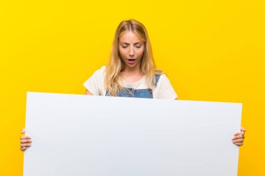 Bir kavram eklemek için boş bir beyaz pankart tutan izole sarı arka plan üzerinde Blonde genç kadın