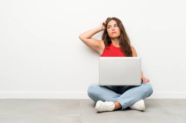 Dizüstü bilgisayarı olan genç bir kadın yerde oturuyor. Şüpheleri var ve kafası karışık.