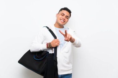 İzole edilmiş beyaz duvardaki sporcu gülümsüyor ve zafer işareti gösteriyor.