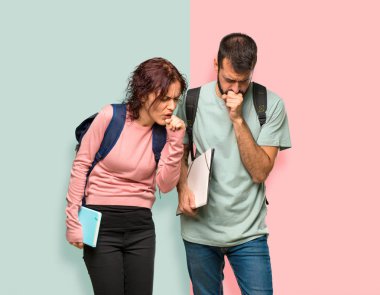 Sırt çantaları ve kitapları olan iki öğrenci öksürük ve renkli duvarda kötü hissediyor.