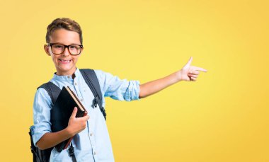 Sırt çantası ve gözlük yan parmak işaret ve sarı arka plan üzerinde bir ürün sunan öğrenci çocuk