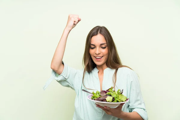 Young woman with salad over isolated green wall celebrating a victory