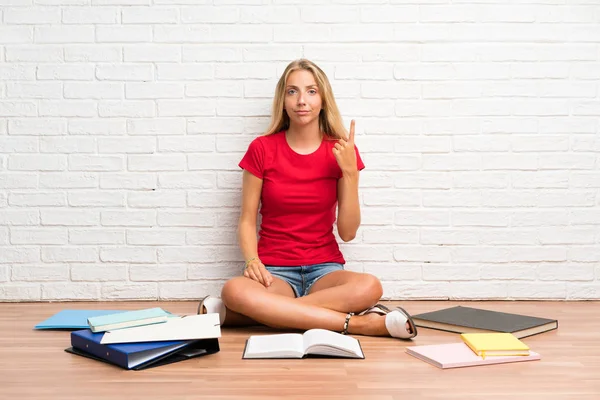 Young blonde student girl with many books on the floor pointing with the index finger a great idea