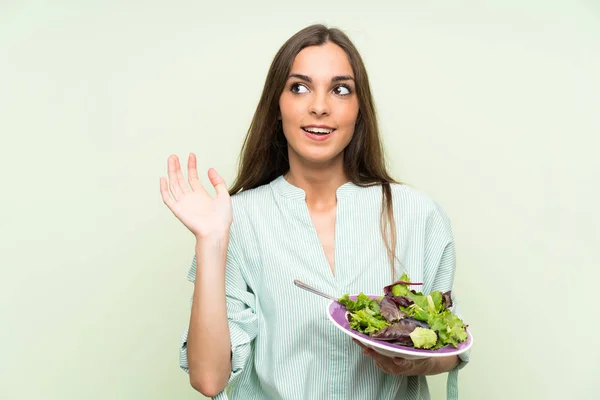 Young woman with salad over isolated green wall saluting with hand with happy expression