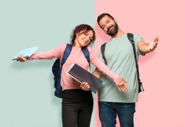 Sırt çantaları ve kitapları sunan ve gelmeye davet eden iki öğrenci. Renkli duvara geldiğiniz için mutlusunuz.