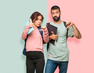 Sırt çantaları ve kitapları olan iki öğrenci iyi-kötü işareti yapıyor. Renkli duvarda evet ya da değil arasında kararsız