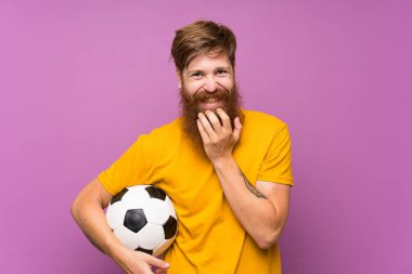 Uzun sakallı kızıl saçlı adam izole mor duvarın üzerinde bir futbol topu tutan çok gülümsüyor