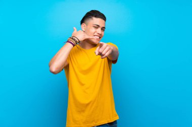 Izole mavi arka plan üzerinde sarı gömlekli genç adam telefon jestyapma ve ön işaret