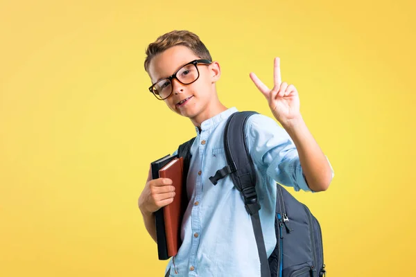 バックパックとメガネを持つ学生の少年が微笑み 黄色の背景に勝利の看板を示しています — ストック写真