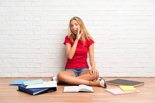 Young blonde student girl with many books on the floor whispering something