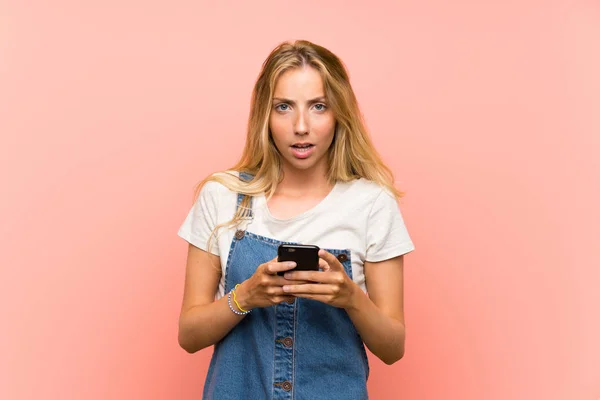 Bionda giovane donna con un telefono cellulare oltre isolato muro rosa — Foto Stock