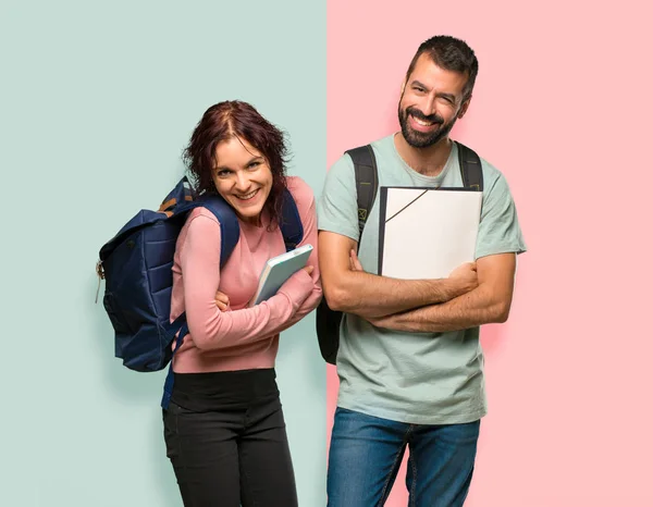 Two students with backpacks and books keeping the arms crossed while smiling on colorful wall