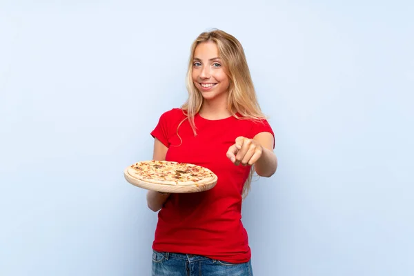 Молодая Блондинка Держит Пиццу Над Изолированной Синей Стеной Указывает Пальцем — стоковое фото