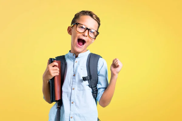 Student boy with backpack and glasses celebrating a victory on yellow background