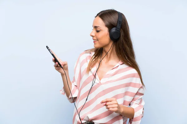 Ung kvinna lyssnar musik med en mobil över isolerad blå vägg — Stockfoto