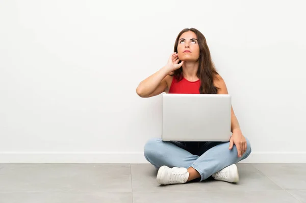 Young woman with a laptop sitting on the floor thinking an idea