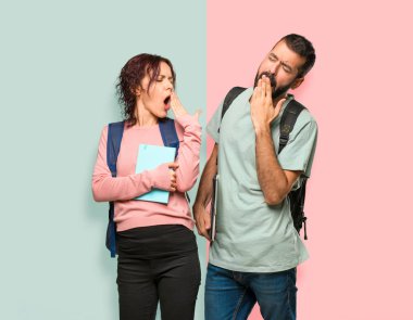 Sırt çantaları ve kitapları esneyen ve el ile ağız kapsayan iki öğrenci. Renkli duvarda uykulu ifade