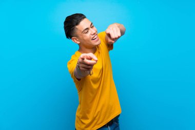 Izole mavi arka plan üzerinde sarı gömlekli genç adam gülümserken size parmak