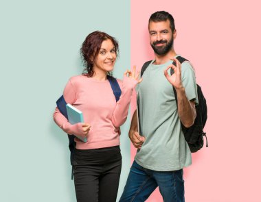 Sırt çantaları ve renkli duvarda parmakları ile ok işareti gösteren kitaplar ile iki öğrenci