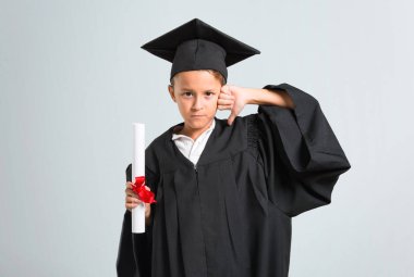 Küçük çocuk gri arka plan üzerinde olumsuz ifade ile başparmak aşağı işareti gösteren mezun