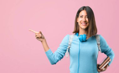 Mavi kazak ve kulaklık ile genç öğrenci kız yan parmak işaret ve pembe arka planda kendine güvenen bir poz gülümseyerek bir ürün sunarken