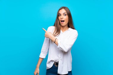 İzole edilmiş mavi arka planı olan genç bir kadın şaşırmış ve işaret ediyor.