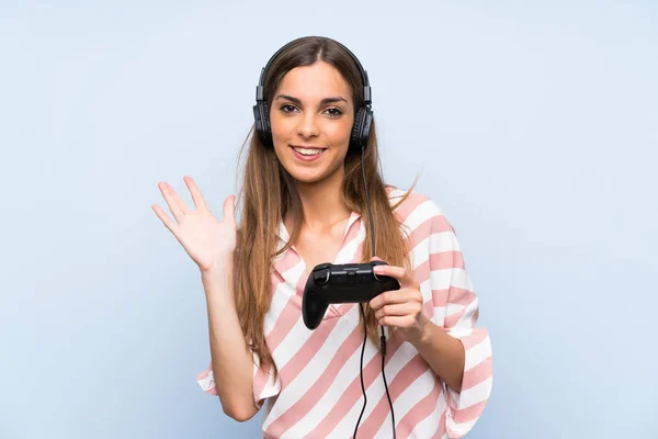年轻女子玩视频游戏控制器在孤立的蓝色墙壁与快乐的表情与手致敬 — 图库照片