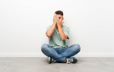 Genç yakışıklı adam gözleri kaplayan ve parmaklardan bakarak katta oturan