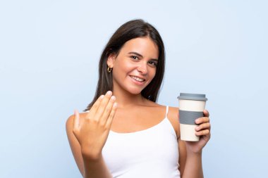 Genç kadın izole mavi arka plan üzerinde bir kahve götürmek tutarak el ile gelmeye davet ediyor. Geldiğin için mutluyum.