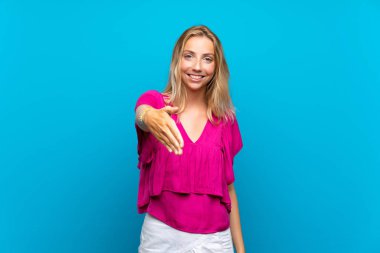 Izole mavi arka plan üzerinde Sarışın genç kadın iyi bir anlaşma kapanış için el sıkışma