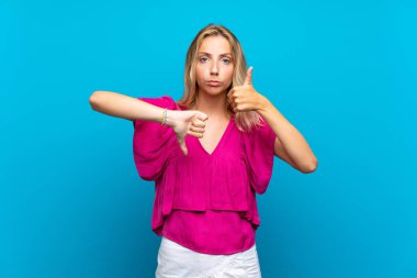 Izole mavi arka plan üzerinde sarışın genç kadın iyi-kötü işareti yapma. Evet ya da değil arasında kararsız