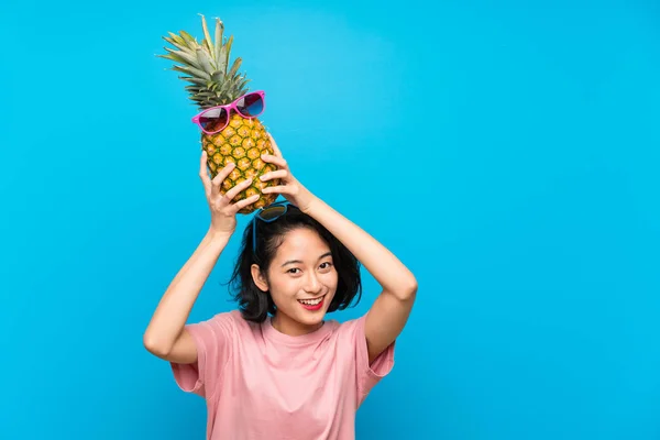 Asiatisk ung kvinna över isolerad blå bakgrund innehar en ananas med sol glasögon — Stockfoto