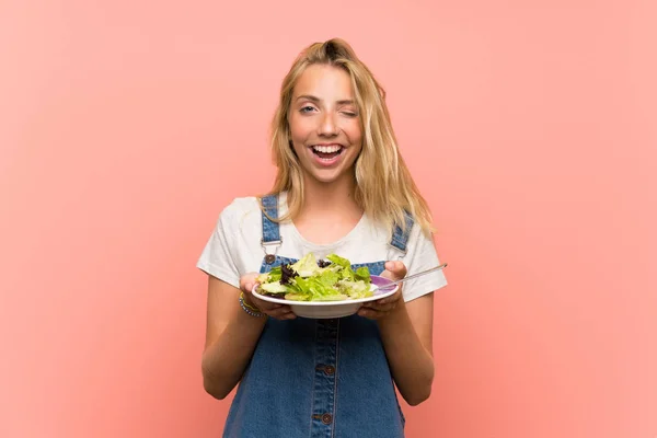 Gelukkige blonde jonge vrouw met salade over geïsoleerde roze muur — Stockfoto