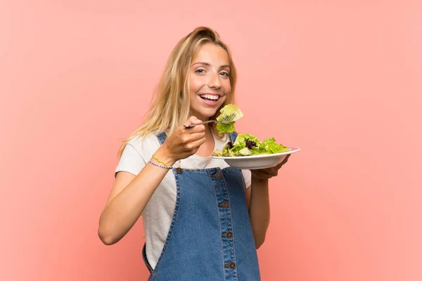 Gelukkige blonde jonge vrouw met salade over geïsoleerde roze muur — Stockfoto