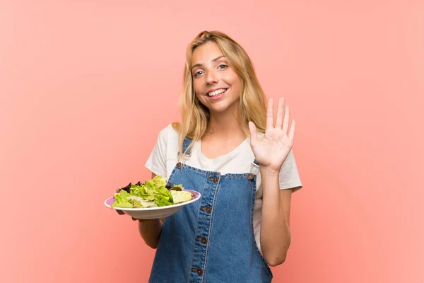 Blonde young woman with salad over isolated pink wall saluting with hand with happy expression