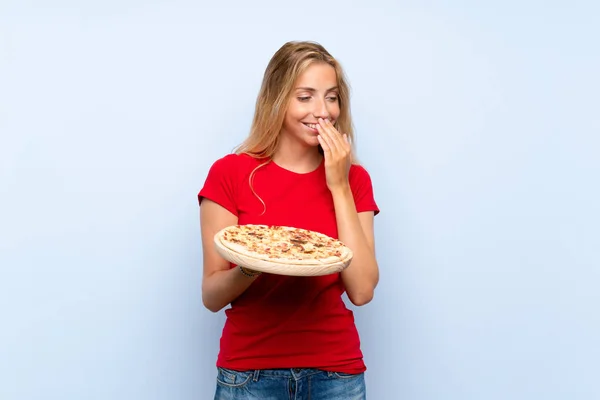 Young blonde woman holding a pizza over isolated blue wall smiling a lot