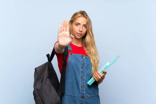 Young blonde student woman over isolated blue wall making stop gesture with her hand