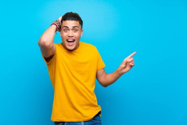 Izole mavi arka plan üzerinde sarı gömlek li genç adam şaşırttı ve yan parmak işaret