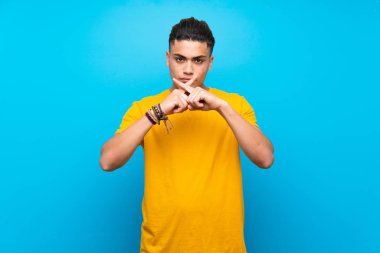 Izole mavi arka plan üzerinde sarı gömlekli genç adam sessizlik jest bir işareti gösteren