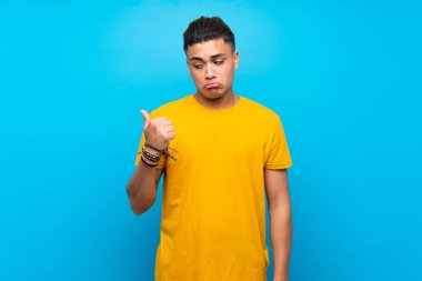 Izole mavi arka plan üzerinde sarı gömlek li genç adam mutsuz ve yan işaret