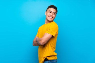 Kolları çapraz ve mutlu izole mavi arka plan üzerinde sarı gömlekli genç adam