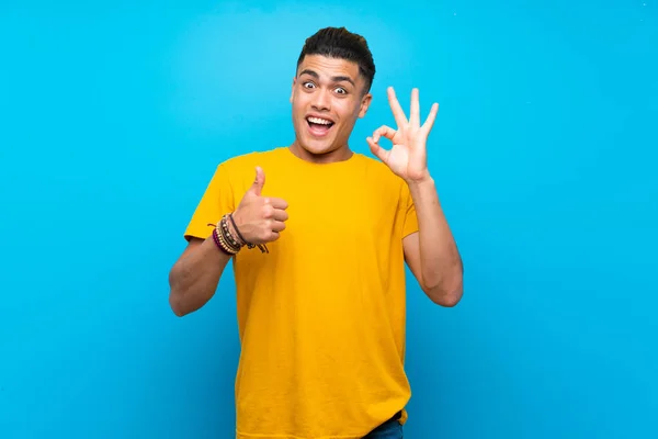 Young man with yellow shirt over isolated blue background showing ok sign and thumb up gesture
